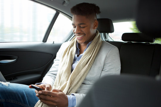 Smiling Young African American Man Looking At Mobile Phone While In Backseat Of Car