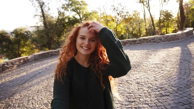 Young caucasian woman in black knitted sweater joyfully running in a colorful autumn park on pavement. Red curly haired girl enjoying autumn foliage, turns around joyfully smiling at camera