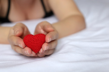 Fototapeta premium Declaration of love, woman holding red knitted heart in hands. Sexy girl in bra lying on the bed, concept of Valentines day, romantic sex, charity, health care or blood donation