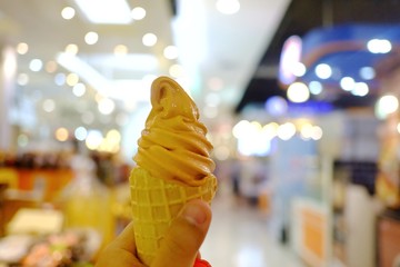 A female hand holding a soft cone of Thai tea ice cream with blur department store area and bokeh light 
