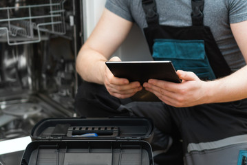 Handyman with tablet pc repairing domestic dishwasher in the kitchen.