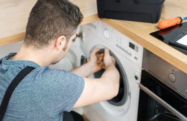 Professional handyman in overalls repairing washing machine in the kitchen.