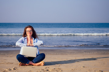 Young calm woman in glasses with laptop freelancing on the tropical paradise beach. Girl freelancer sitting in a summer sand on the seashore dream remote work indian ocean background copyspace