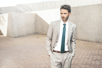 Young attractive serious businessman is standing next to a concrete wall background wearing light classic tuxedo. Urban working style.