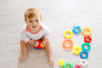 Fototapeta na wymiar Adorable baby girl playing with educational toys in nursery. Happy healthy child having fun with colorful different toys at home. Kid trying to build plastic pyramid and using blocks with letters
