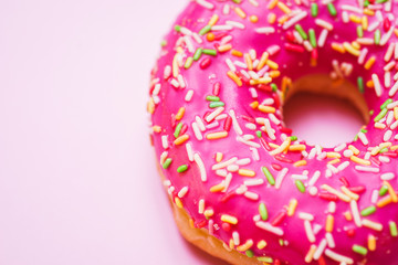 Bright donut in a pink glaze with a multi-colored rainbow sprinkle on a pink background. 