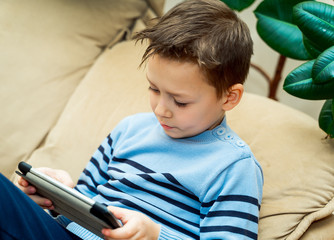 Little boy sitting on light couch and touching the screen of wireless tablet at home. Portrait of a young child with a laptop.