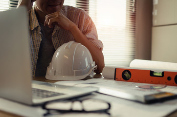 young asian engineer, architect, contractor thinking and worrying about new project contract plan with laptop and white safety helmet on desk in office center at construction site, contractor concept
