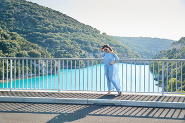 The beautiful girl in a blue dress and sunglasses poses on the bridge, the long chestnut hair,...