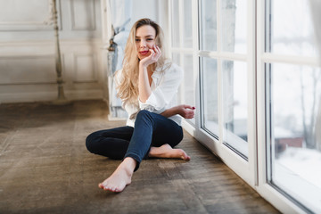 Obraz premium Model blonde white shirt blue pants jeans sitting on wooden parquet floor by window.
