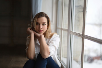 Model blonde white shirt blue pants jeans sitting on wooden parquet floor by window.