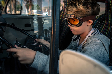 Blond child driving a vintage camper wearing aviator goggles sitting on colorful seat, puts his...