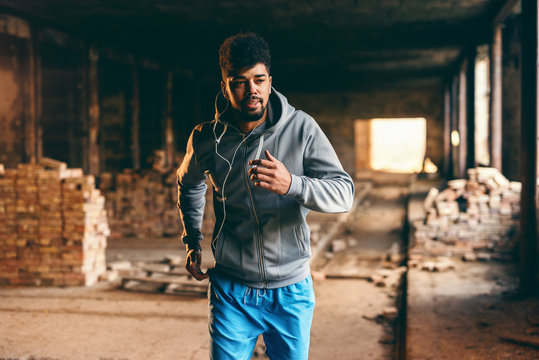 Young sporty african american guy in sportswear and with earphones running in old brick factory.