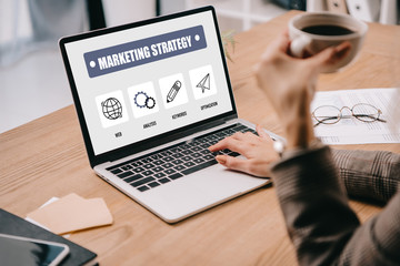 cropped view of businesswoman with coffee cup typing on laptop with marketing strategy