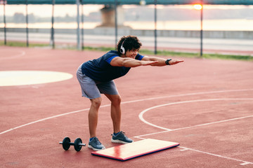 Obraz premium Sporty man with curly hair doing exercises on the court and having earphones in ears. Summertime in the morning.