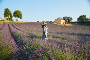 The beautiful young girl in a blue dress walks across the field of a lavender, long curly hair, smile, pleasure, mountains on background, a house of the gardener, trees, perspective of a lavender