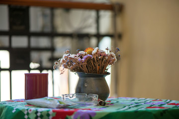 Vase of beautiful dried flowers standing on table with colorful tablecloth next to cigarettes, ash tray and lighter. Concept of unhealthy morning habits.  