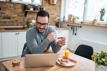 Young man sitting at the kitchen table with breakfast in morning using laptop and credit card.