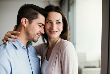 Young couple in love standing by the window at home, hugging. Copy space.