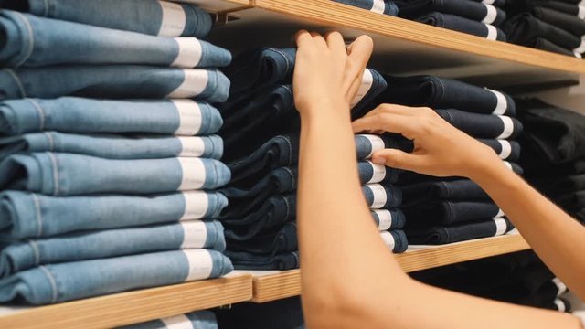 Young Woman Taking Pair of Blue Denim Jeans From Stacks in Clothing Shop. Female Hands Choosing Correct Pants Size in Shopping Mall. 4K Slowmotion Consumerism Concept.