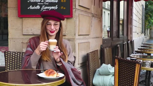 Beautiful girl with long hair, red lipstick, glasses, beret sitting having breakfast at the french cafe, drink coffee with croissant and smiling
