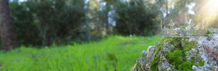 mysterious and magical photo of silver king crown over the stone covered with moss in the England woods or field landscape with light flare. Medieval period concept.