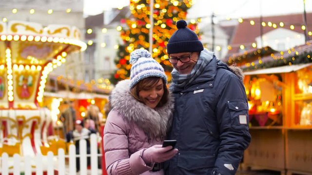 technology, winter holidays and people concept - happy senior couple taking selfie by smartphone at christmas market on town hall square in tallinn, estonia