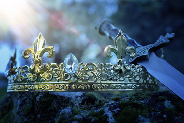 mysterious and magical photo of silver king crown and sword over the stone covered with moss in the...