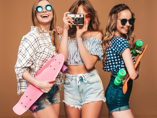 Three beautiful stylish smiling girls with colorful penny skateboards.Women in summer hipster checkered shirt clothes posing on golden background.Taking pictures on retro camera