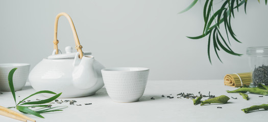 Asian tea concept, two white cups of tea and teapot surrounded with green tea dry leaves , space for a text on white background. Brewing and Drinking tea.  long, wide banner