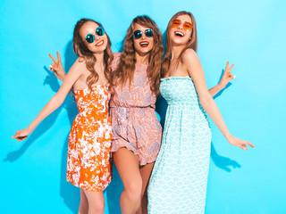 Three young beautiful smiling hipster girls in trendy summer casual dresses and in sunglasses. Sexy carefree women posing near blue wall. Positive models going crazy