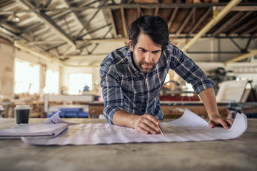 Woodworker leaning on a bench in his workshop reading blueprints