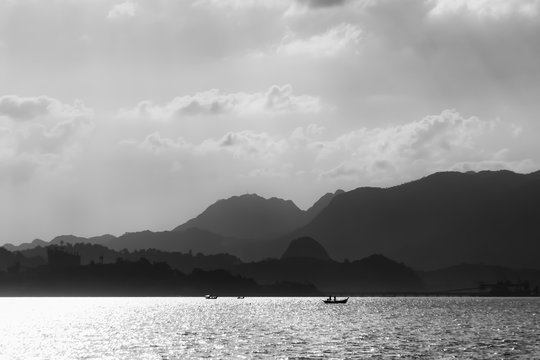 Silhouettes of fishing boats against the backdrop of mountains and a sunny sky with clouds. Black and white photography