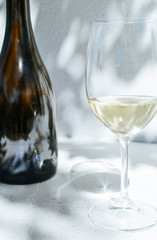 White wine bottle and a glass standing in front of a white concrete wall with shadows of leaves on a sunny summer day 