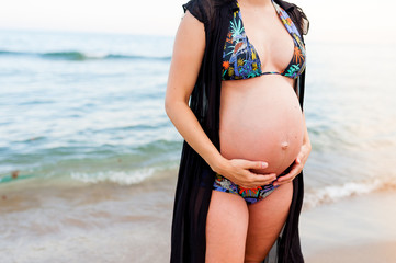 Fototapeta na wymiar front close up of big belly of young pregnant woman close to the ocean during moody autmn day in color swimsuit with black dress