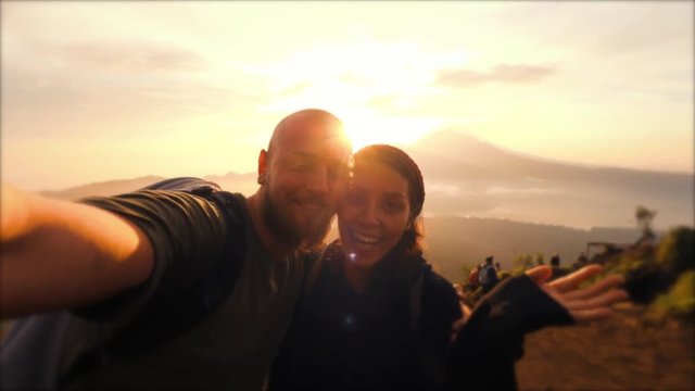 Young Mixed Race Couple Taking Selfie Portrait on Montain Peak with Beautiful Sunrise Landscape on Background. 4K Slowmotion Lifestyle Hiking Travel Concept Footage. Batur Volcano, Bali, Indonesia.