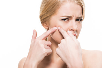 unhappy young woman squeezing pimple on face and looking at camera isolated on white