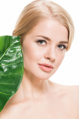 attractive naked girl with green tropical leaf near face looking at camera isolated on white