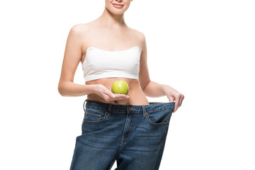 cropped shot of smiling young woman in oversized jeans holding green apple isolated on white