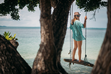 A young woman is swinging on a swing in the shade of a big tree by the sea. Girl on the beach swing on the coast of a tropical island.