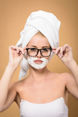 woman with towel on head and shaving cream on face adjusting eyeglasses and looking at camera isolated on beige