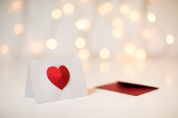 A white greeting card with a red 3d heart for Valentine's day next to a red envelope on a fairy light bokeh background