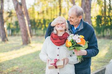 Happy elderly man gives his lady flowers while covering her eyes with palm, she have some present for him - Valentine’s Day celebrating, love and getting old together concept.