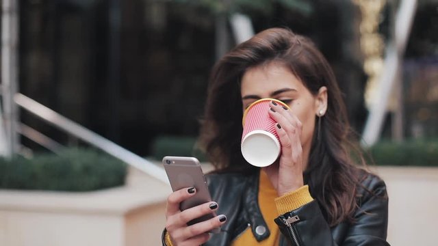 Young happy woman using smartphone and drinking coffee to go outdoors, walking down the city street, slow motion