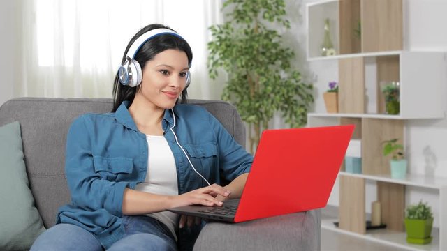 Happy woman wearing headphones browsing laptop content sitting on a couch at home