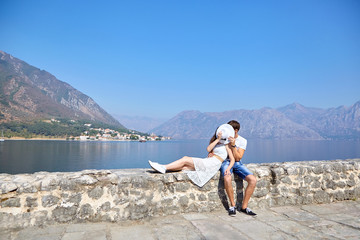 Cute couple in love kissing behind white female hat , sitting on the stones on the background of the sea coast, mountains and blue sky