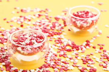 Colorful heart shaped sprinkles in glass bowls on yellow background