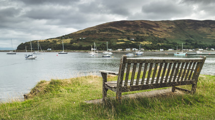 Close-up bench on the Arran island shore. Amazing bayview. Breathtaking Scottish landscape. Cozy bay with the yachts under the rainy cloudy sky. Ideal place for the rest and relax.