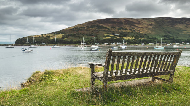 Fototapeta Close-up bench on the Arran island shore. Amazing bayview. Breathtaking Scottish landscape. Cozy bay with the yachts under the rainy cloudy sky. Ideal place for the rest and relax.
