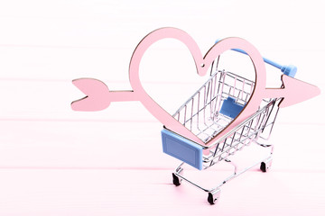 Shopping cart and wooden heart with arrow on pink table